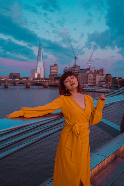 A woman wearing the Sarvin Mustard Wrap Maxi Dress with balloon bishop sleeves stands by a bridge railing at dusk, city skyline and river behind her. The partly cloudy sky glows in blue and purple hues, cranes visible in the distance.