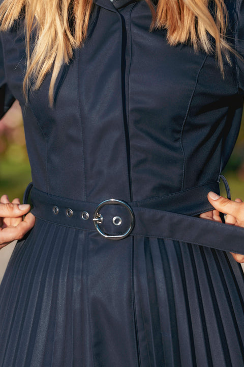 Blue pleated coat with a belt being adjusted against a blurred green background.