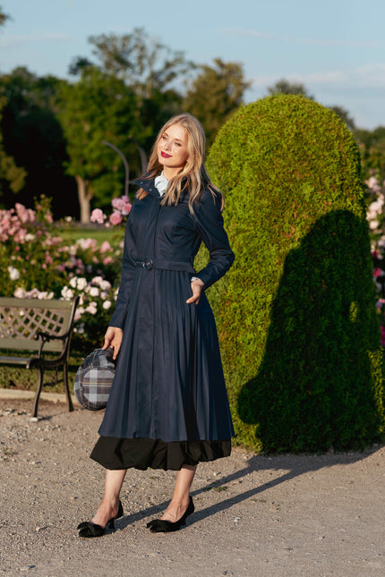 Woman in a dark blue coatstanding in a garden with greenery and flowers.