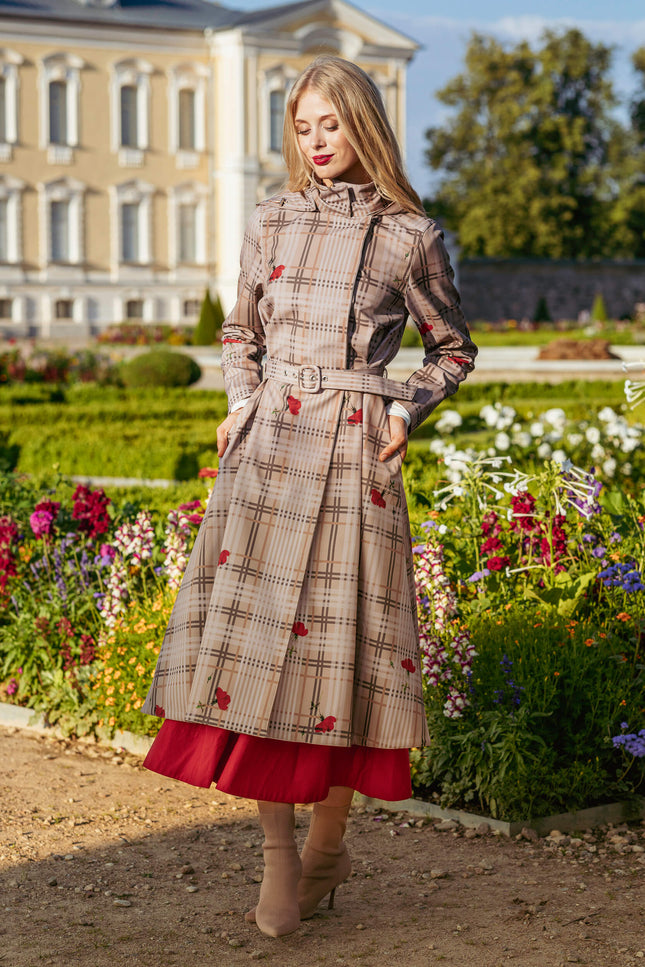 Woman in a plaid beige color coat with red accents standing in a garden with a classical building in the background