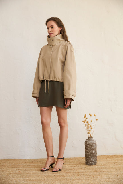 A model wearing a beige mini woolen skirt with dual side zippers, paired with a light-colored, long-sleeved top and standing next to a decorative vase.