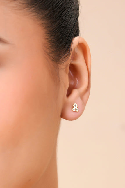 A close-up image of a person's ear wearing a single gold-colored stud earring with a triple crystal bezel.