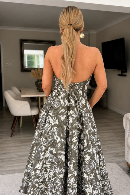 A woman with long blonde hair in a low ponytail stands indoors, facing away, wearing the Sarvin Tala Corset Midi Dress—a strapless black, white, and gray floral piece—in a modern living and dining area.
