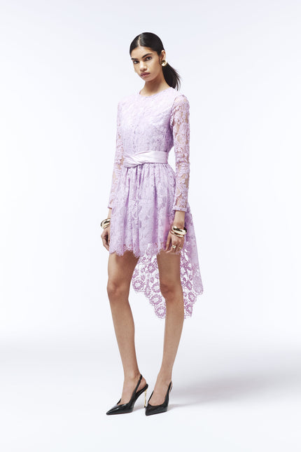 Romantic high-low lace dress in pastel lilac with draped satin belt and scalloped hem.