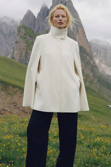 Person wearing a white coat standing in a mountainous landscape with green grass and yellow flowers.