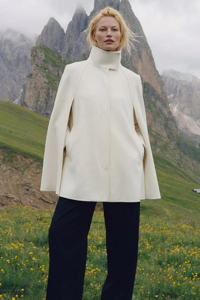 Person wearing a white coat standing in a mountainous landscape with green grass and yellow flowers.