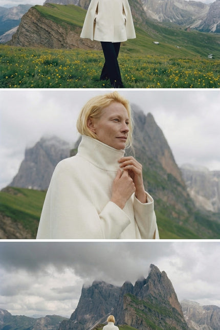 Woman in a white coat standing in a mountainous landscape with green fields and rocky peaks.