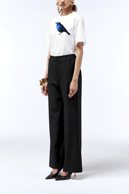Model wearing the white starling print T-shirt untucked with black trousers, pointed heels and gold jewellery.