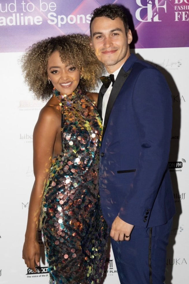 A woman wearing the Sarvin High Neck Backless Sequin Mini Dress poses and smiles next to a man in a blue suit and bow tie, both standing in front of an event step-and-repeat backdrop.