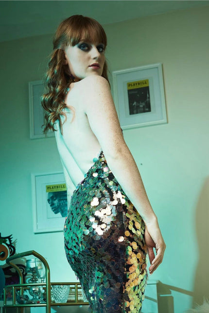 A person with long red hair stands in a room, wearing the Sarvin High Neck Backless Sequin Mini Dress. Framed Playbill posters are on the wall behind them, illuminated by greenish-blue lighting.