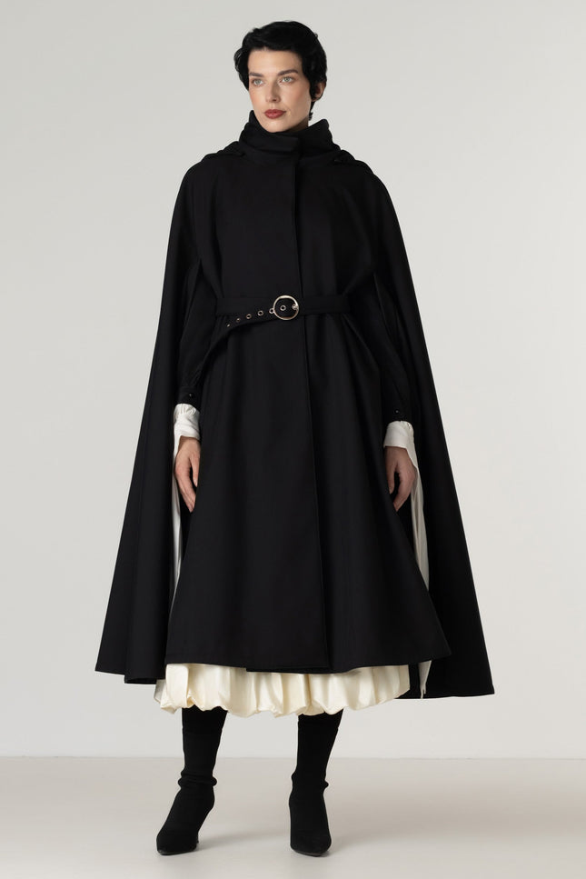A model wearing a long black cape-style coat by RainSisters with a high mock neck, a matching waist belt with a round metal buckle, and layered white ruffled cuffs and hem, paired with black boots against a light gray studio background.