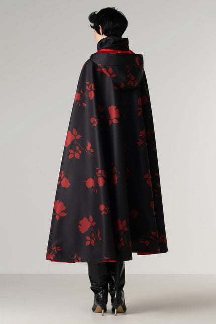 A model viewed from the back wearing a black hooded cape with a red floral pattern and a red inner lining, paired with black boots, against a light gray studio background.