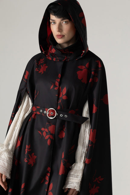 A model wearing a black hooded cloak by RainSisters with a red floral pattern, styled with sheer white ruffled blouse and a black belt with a silver buckle.