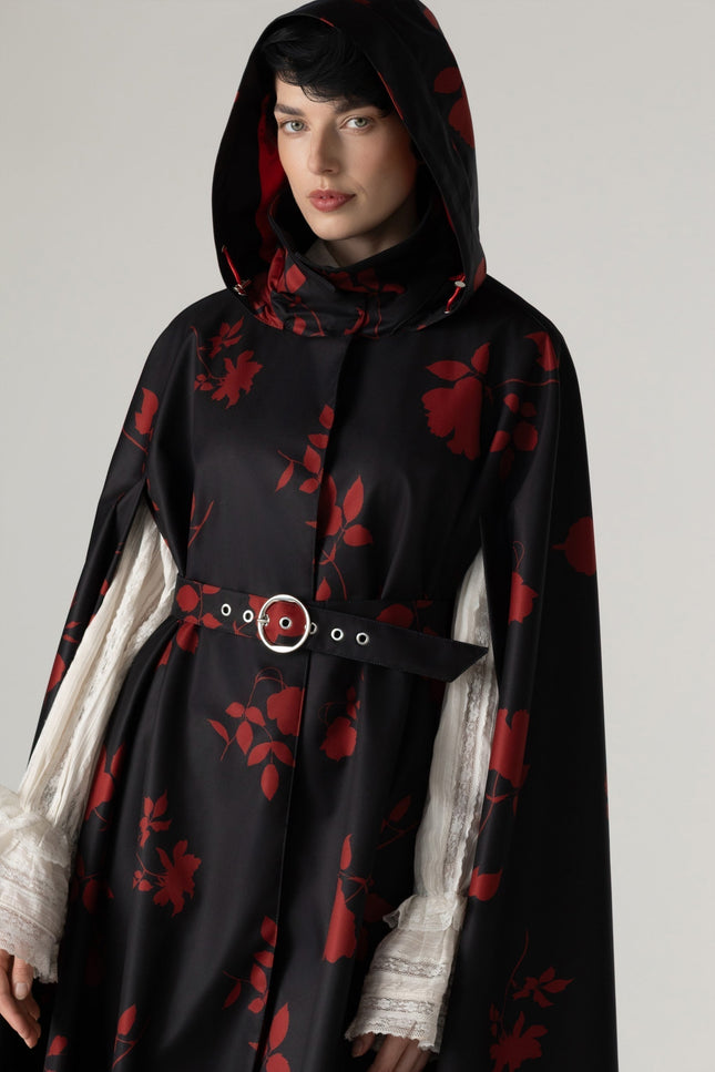 A model wearing a black hooded cloak by RainSisters with a red floral pattern, styled with sheer white ruffled blouse and a black belt with a silver buckle.