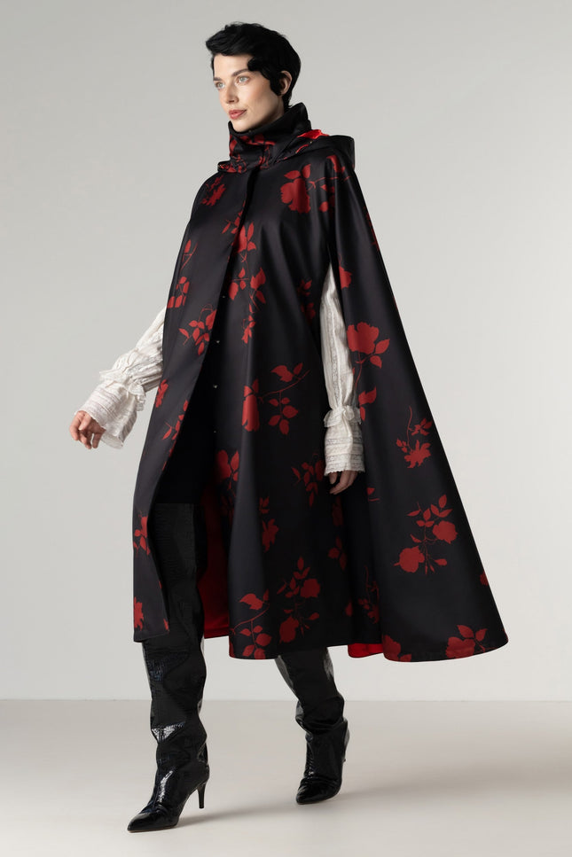 A model wearing a long black cape with a red rose pattern by RainSisters, styled over a white ruffled shirt, black pants, and black high-heeled boots, set against a plain light gray studio background.