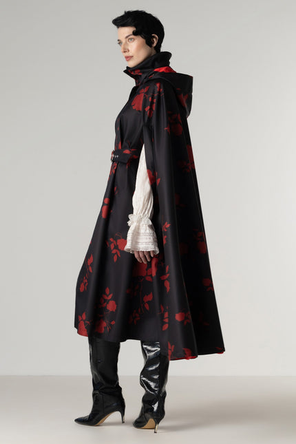 A model is shown from the side wearing a long black hooded cape with a red floral pattern by RainSisters, styled over a white ruffled long-sleeve blouse, black pants, and black high-heeled boots against a light gray studio background.