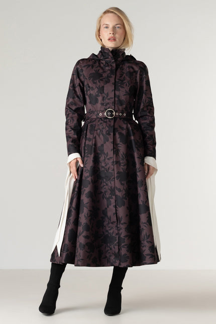 A model wears a long, high-neck coat with a dark floral pattern in black and deep brown and a matching belt with a buckle.