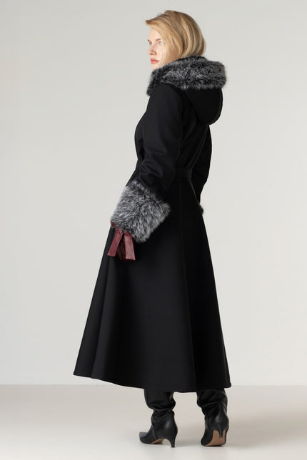 A woman modeling a long black coat with a gray faux-fur trimmed hood and matching sleeve fur, paired with black heeled boots against a plain light gray studio background.