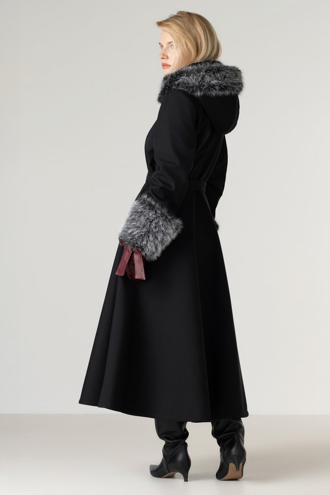 A woman modeling a long black coat with a gray faux-fur trimmed hood and matching sleeve fur, paired with black heeled boots against a plain light gray studio background.