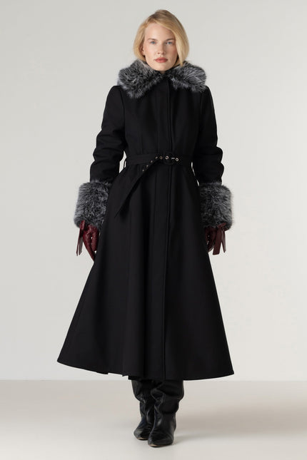 A woman modeling a long black coat with a gray fur-trimmed collar and matching gray fur-trimmed cuffs, wearing burgundy gloves and black boots against a plain light gray studio background.