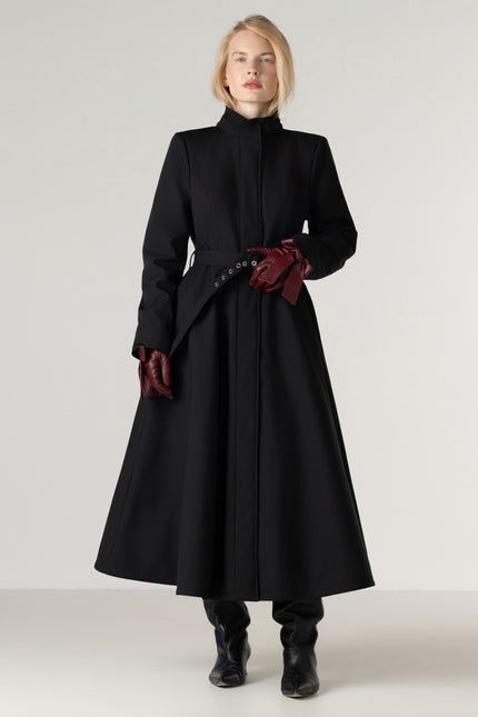 A model wearing a long black high-neck coat with a matching waist belt and dark red gloves, standing against a light gray studio background.