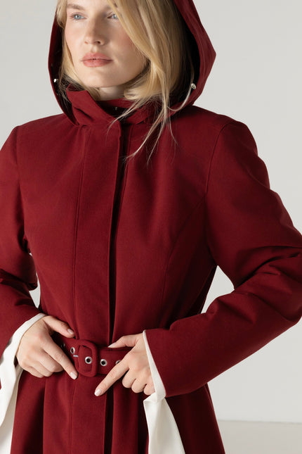 A woman wearing a deep burgundy hooded coat with a matching belt, photographed against a plain light gray studio background.