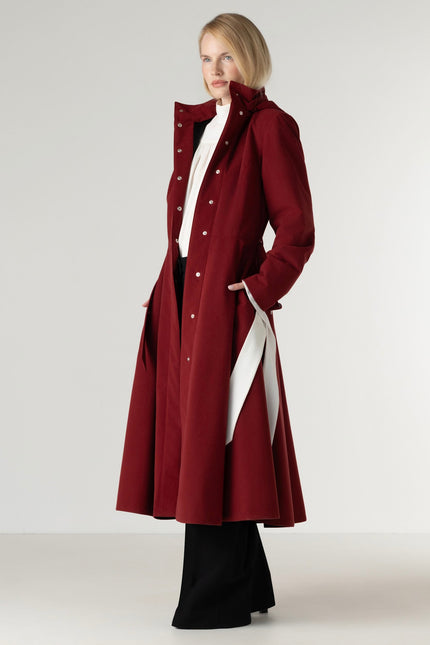A woman modeling a long burgundy coat with silver-tone buttons, worn over a white top and black wide-leg pants, against a plain light gray studio background.