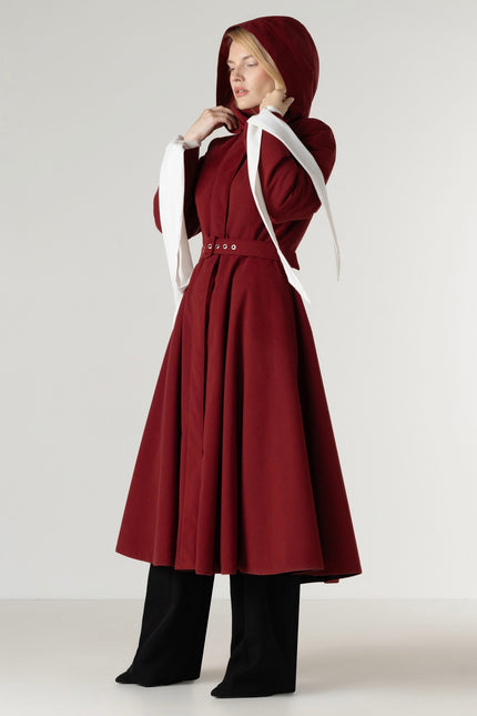 A woman models a long burgundy hooded coat with a matching waist belt, styled with black wide-leg pants against a light gray studio background.