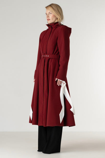 A model wearing a long, high-neck, deep burgundy coat with a matching waist belt, paired with black wide-leg trousers, against a plain light gray studio background.