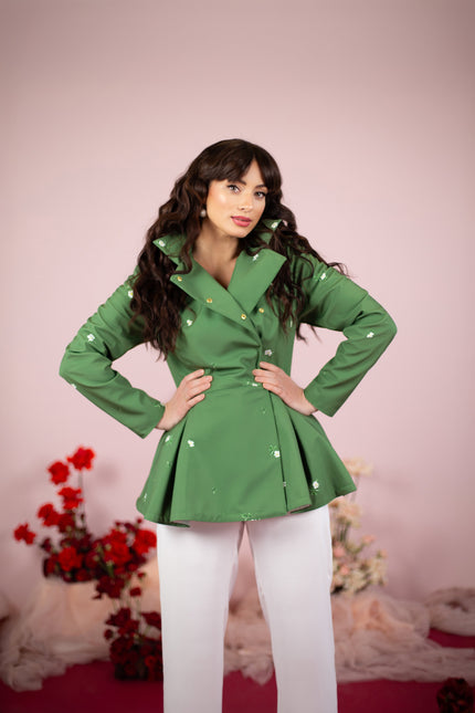 A model wearing a double-breasted green jacket with a detachable hood and white floral print, paired with white trousers, standing against a pink background with floral decorations.