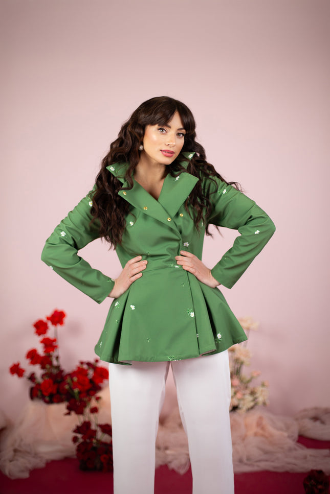 A model wearing a double-breasted green jacket with a detachable hood and white floral print, paired with white trousers, standing against a pink background with floral decorations.