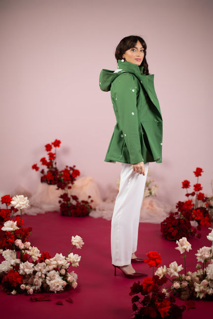 Woman in a green coat standing in front of floral decorations on a pink background