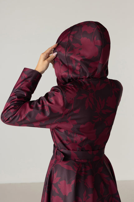 A person shown from the back wearing a hooded long coat with a dark floral pattern in deep burgundy and dark brown by RainSisters, against a plain light gray studio background.