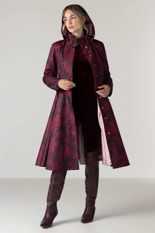 A woman models a long hooded coat with a dark red and black abstract pattern by RainSisters, worn over a dark dress and brown over-the-knee boots against a light gray studio background.