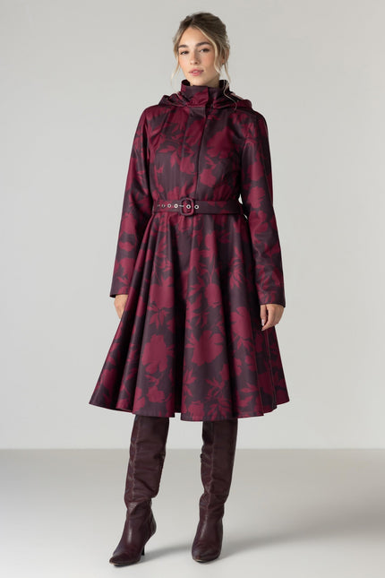 A woman models a long hooded coat with a dark red and black floral pattern by RainSisters, featuring a belted waist and a flared skirt.
