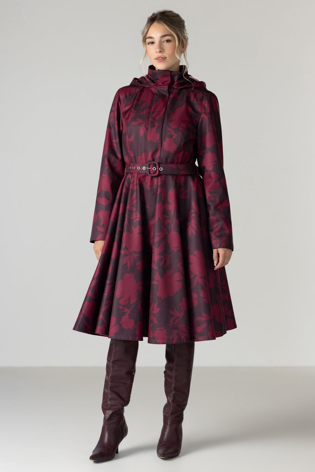 A woman models a long hooded coat with a dark red and black floral pattern by RainSisters, featuring a belted waist and a flared skirt.