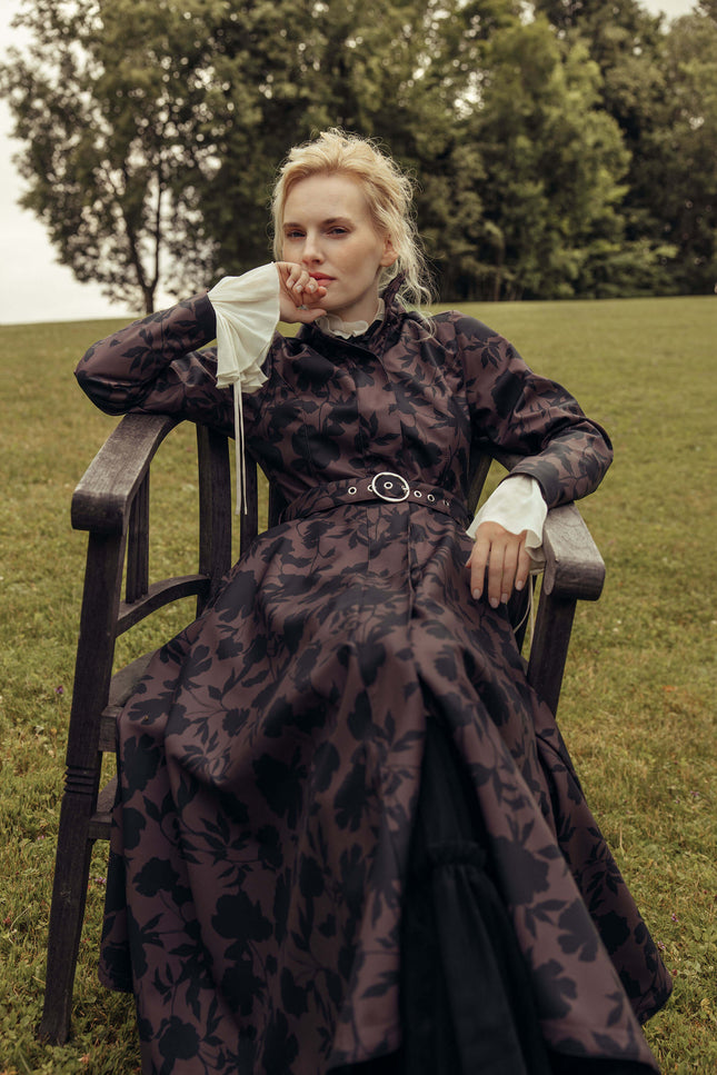 Model sitting outdoors wearing the Women's Brown Floral Fit and Flare Coat Brocade Dusk, showcasing the waterproof softshell fabric, elegant floral print, and timeless feminine silhouette.