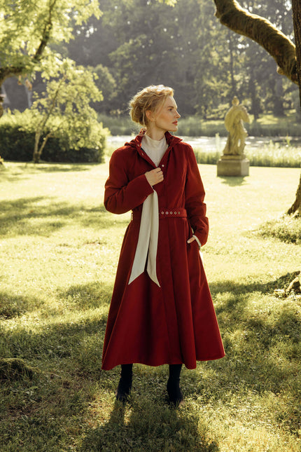 A woman stands in a sunlit park wearing a long red coat over a white top and black boots. Trees, grass, and a statue are visible in the background.