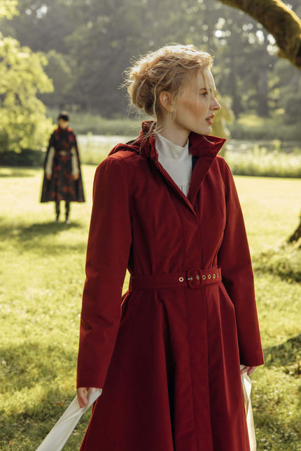 A woman wearing a long burgundy belted coat over a white top stands in a sunlit grassy park. Another person in a dark patterned coat is visible in the blurred background.
