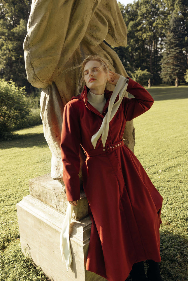 A woman in a long red coat leans against a stone statue in a grassy park setting.