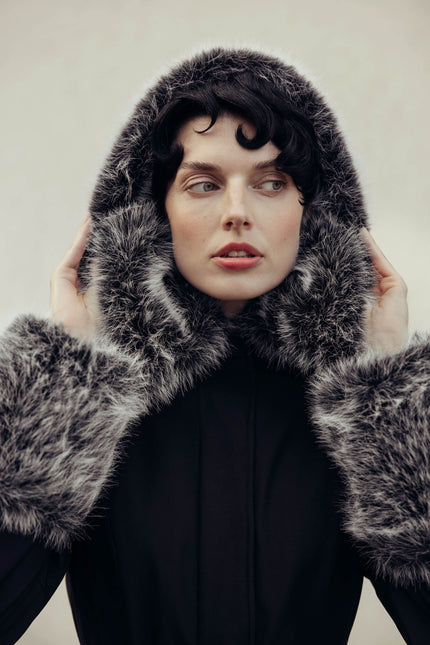 A model wearing a luxurious black and gray fur hooded coat with faux-fur trim, set against a plain light beige background.