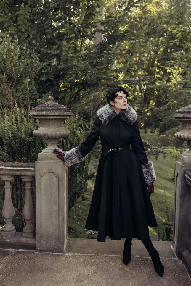 A woman wearing a long black coat with a gray fur collar and matching gloves stands on a stone terrace in a garden setting.