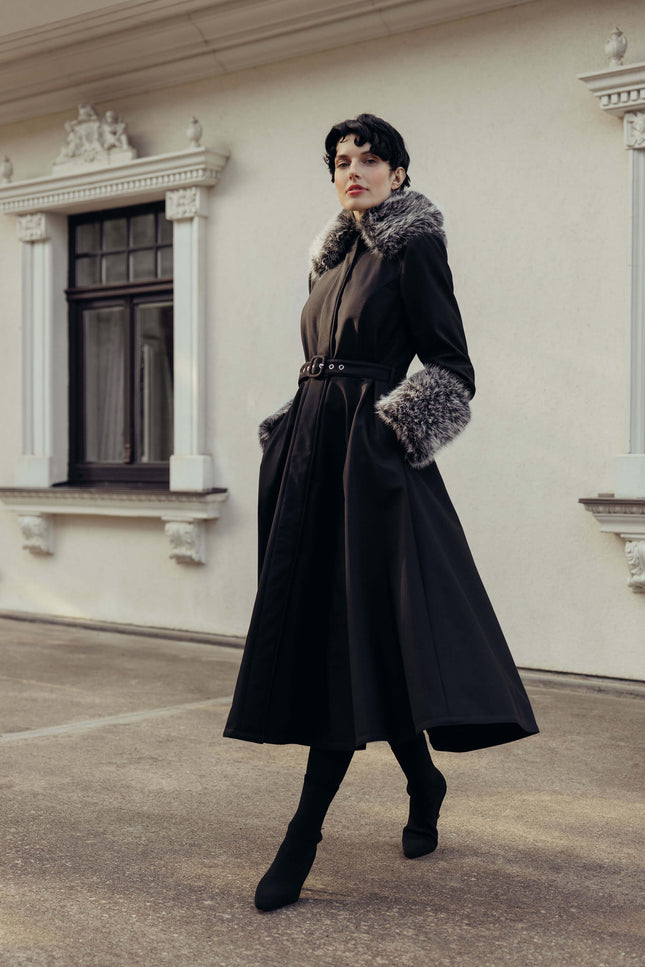 A woman models a long black winter coat with a gray and white fur-trimmed collar and cuffs, paired with a matching black belt and black boots, standing in front of a cream-colored building.