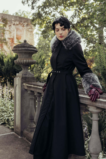Outdoor full-length view of a women's black winter coat with detachable faux fur, featuring a fitted bodice, flowing skirt, and matching buckle belt.