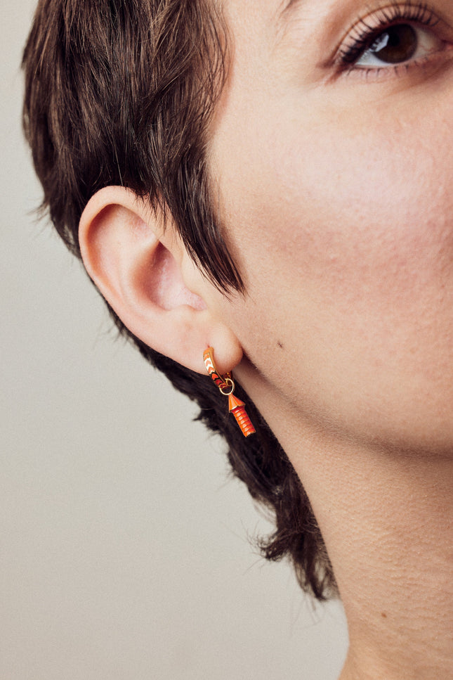 image of rocket enamel earrings in orange and gold shown close-up of ear and eye on model with white skin and short brown hair