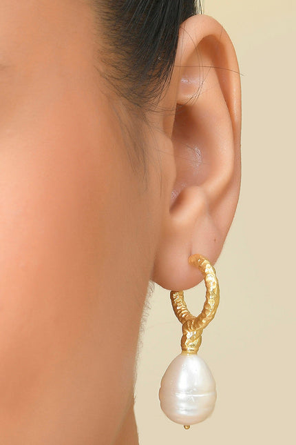 A close-up image of a woman's ear wearing a gold-colored drop hoop earring with a white pearl.