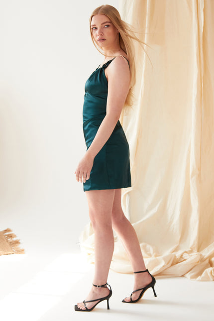 A woman with long blonde hair stands in profile wearing the Sarvin Emerald Green Silk Satin Backless Mini Dress and black strappy heels, indoors before a beige fabric backdrop in soft lighting.