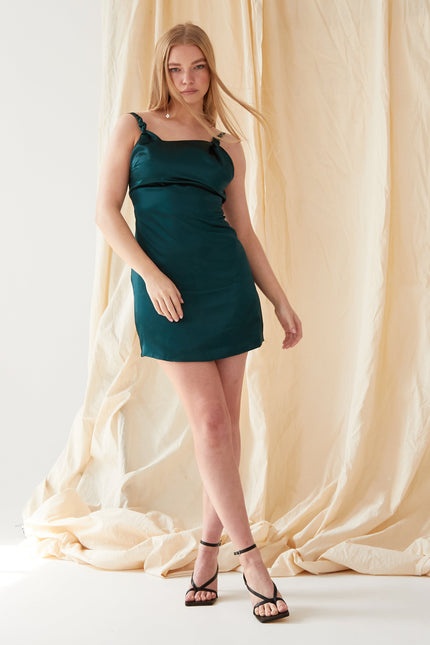A woman with straight blonde hair and a neutral expression stands against a beige fabric backdrop, wearing the Sarvin Emerald Green Silk Satin Backless Mini Dress and black strappy heels, with one leg slightly forward.
