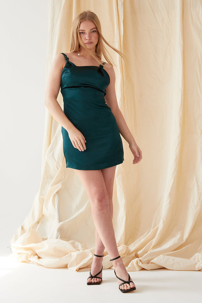 A woman with straight blonde hair and a neutral expression stands against a beige fabric backdrop, wearing the Sarvin Emerald Green Silk Satin Backless Mini Dress and black strappy heels, with one leg slightly forward.
