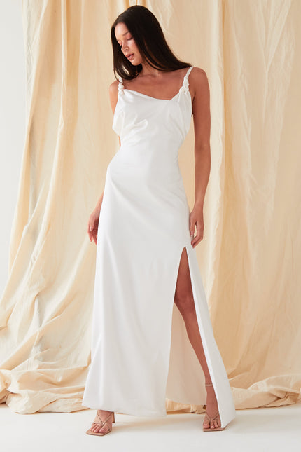 A woman in the Sarvin Ivory Satin Asymmetric Backless Maxi Dress stands against a beige fabric backdrop, wearing strappy heels, her hair down, and gazing downward with effortless elegance.